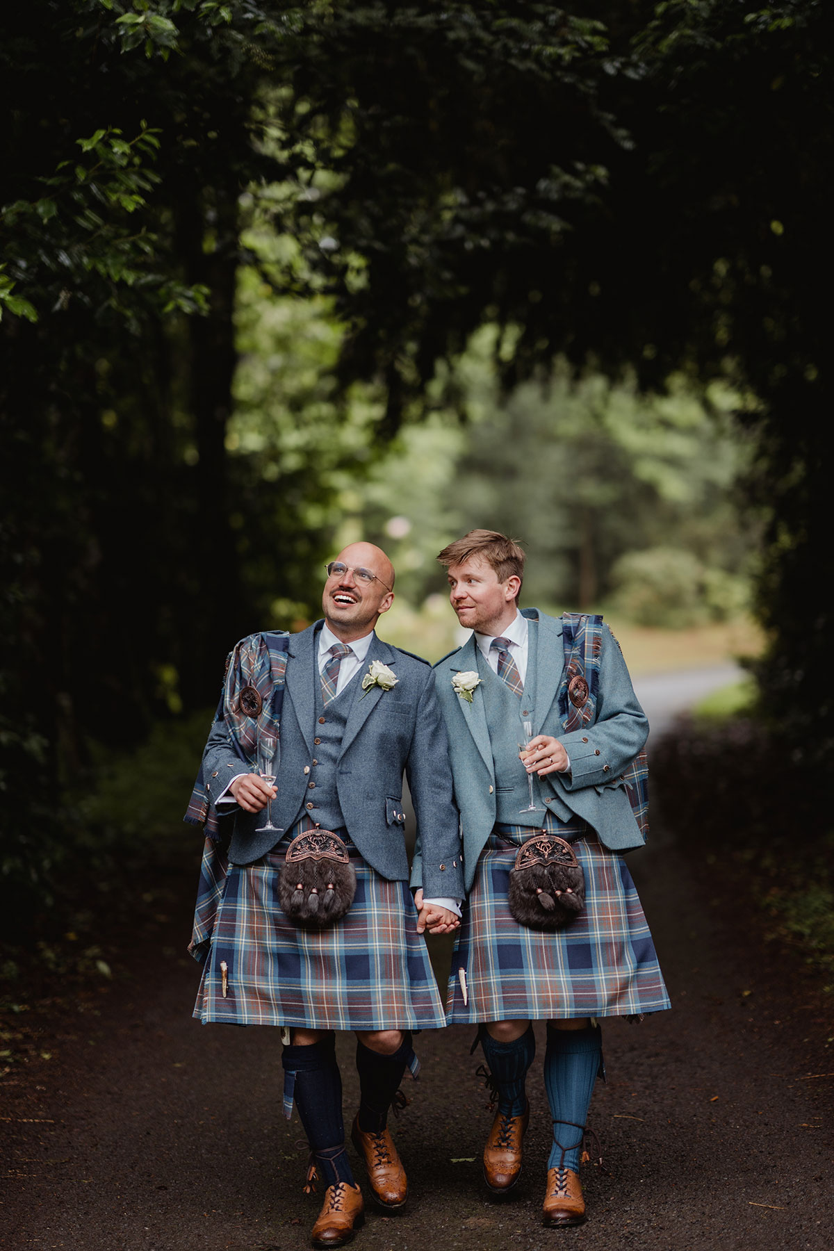 The couple walk hand in hand along a tree-lined path, smiling and chatting while dressed in coordinating blue tartan kilts