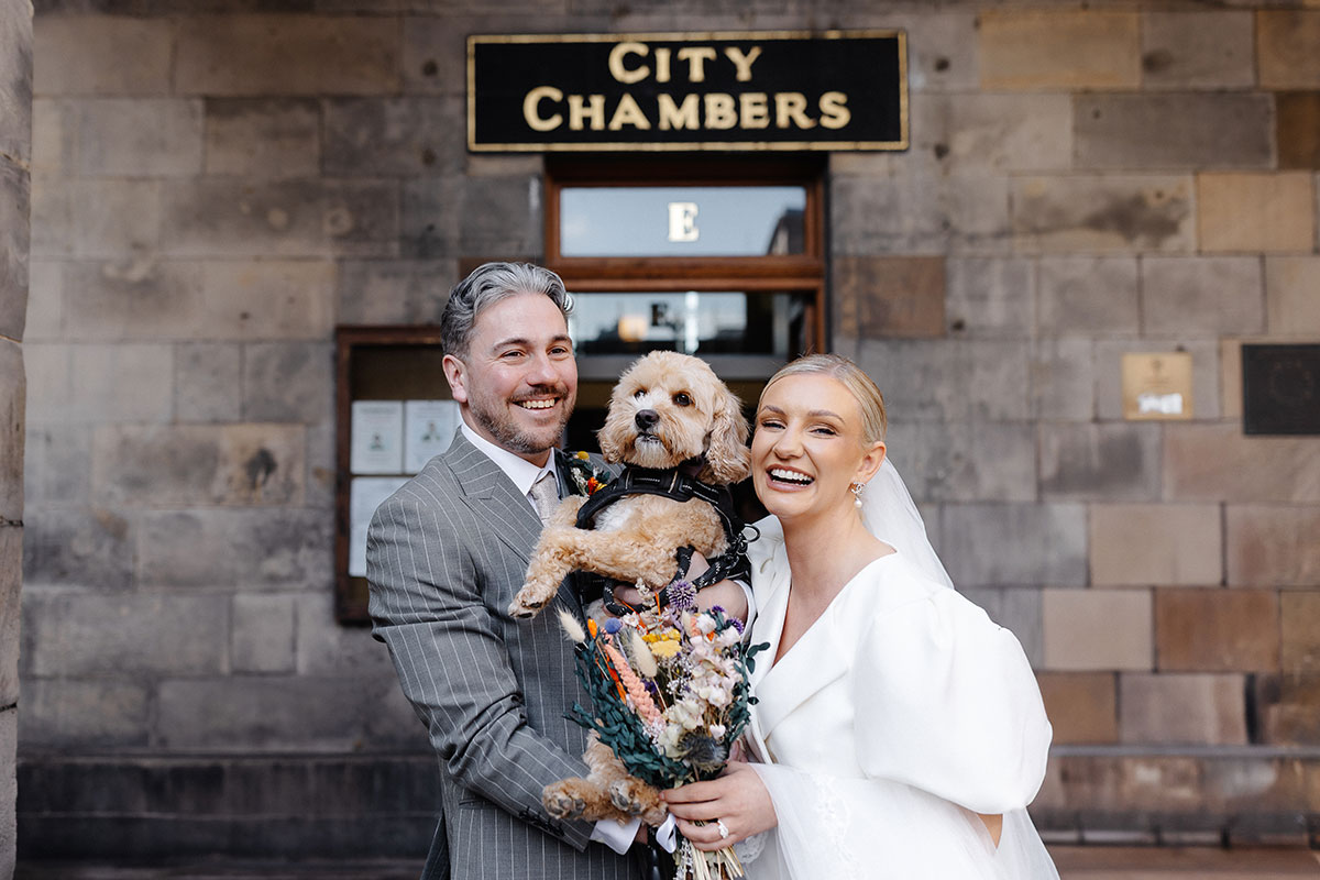 Bride and groom pose with their dog outside Edinburgh City Chambers after their registry office wedding, smiling beneath the historic stone archway.