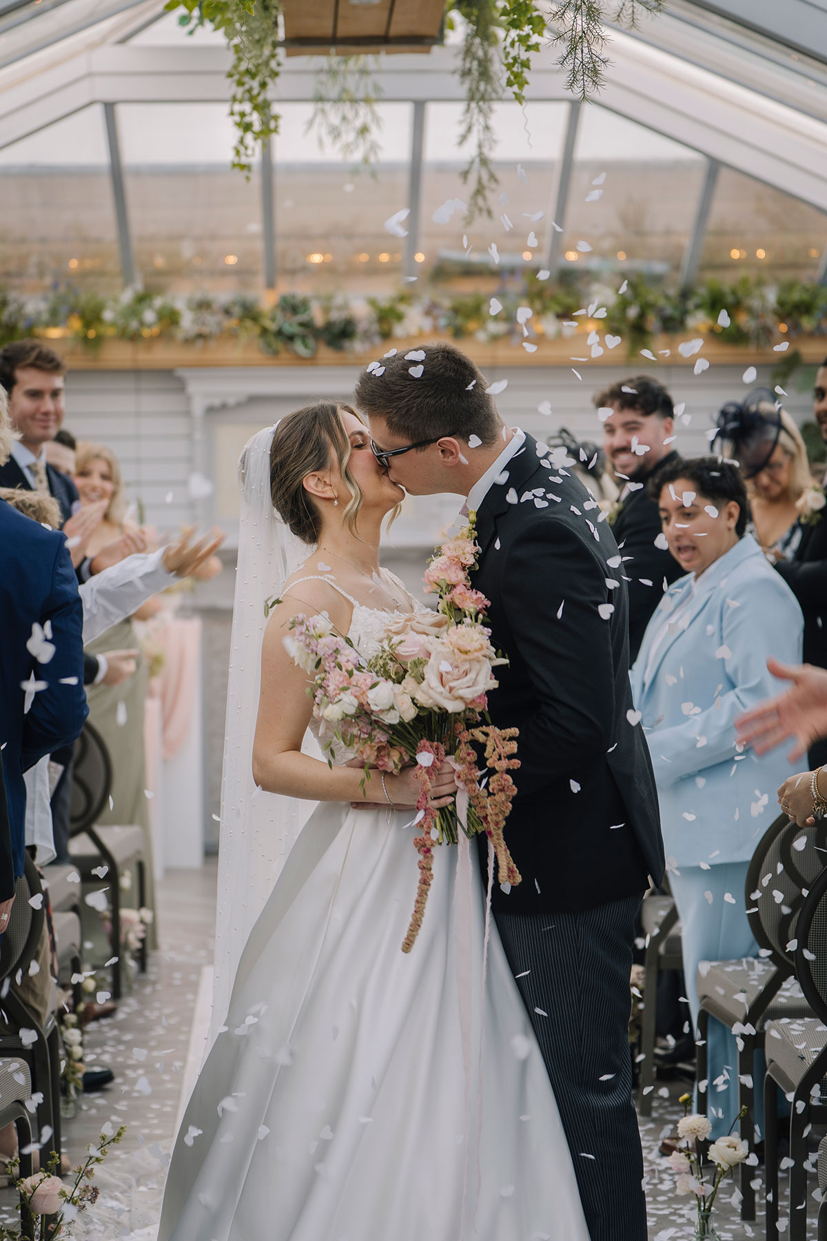Bride and groom kissing under falling confetti after their wedding ceremony while guests celebrate around them
