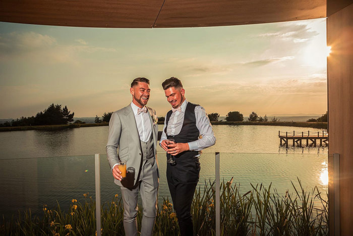 two grooms standing on a glass-fronted terrace at sunset at The Vu, a sustainable waterside wedding venue in Scotland