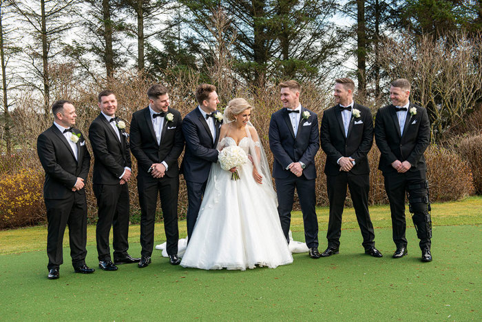 A Bride Poses With Seven Men Wearing Black Dinner Suits On Grass