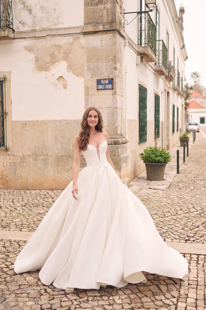 brunette bride stands smiling in cobbled street wearing a strapless ballgown wedding dress with a plunging illusion neckline