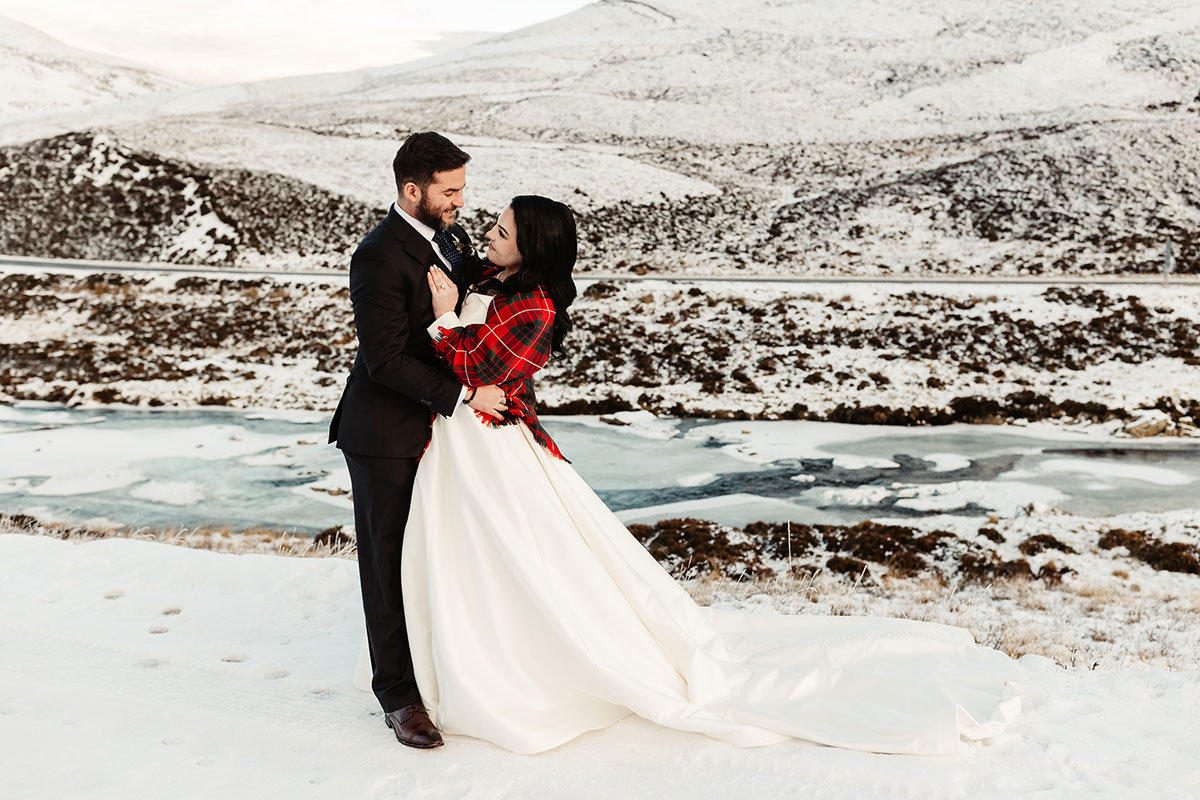 Bride wrapped in a red tartan shawl as she embraces the groom on a snowy hillside above a frozen river