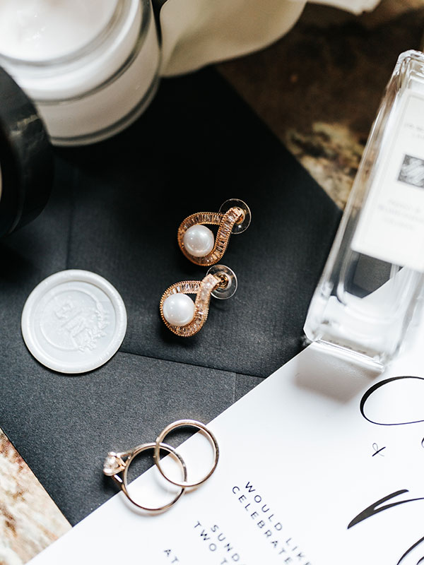 Close-up of wedding rings and pearl earrings styled with invitation stationery and bridal perfume details