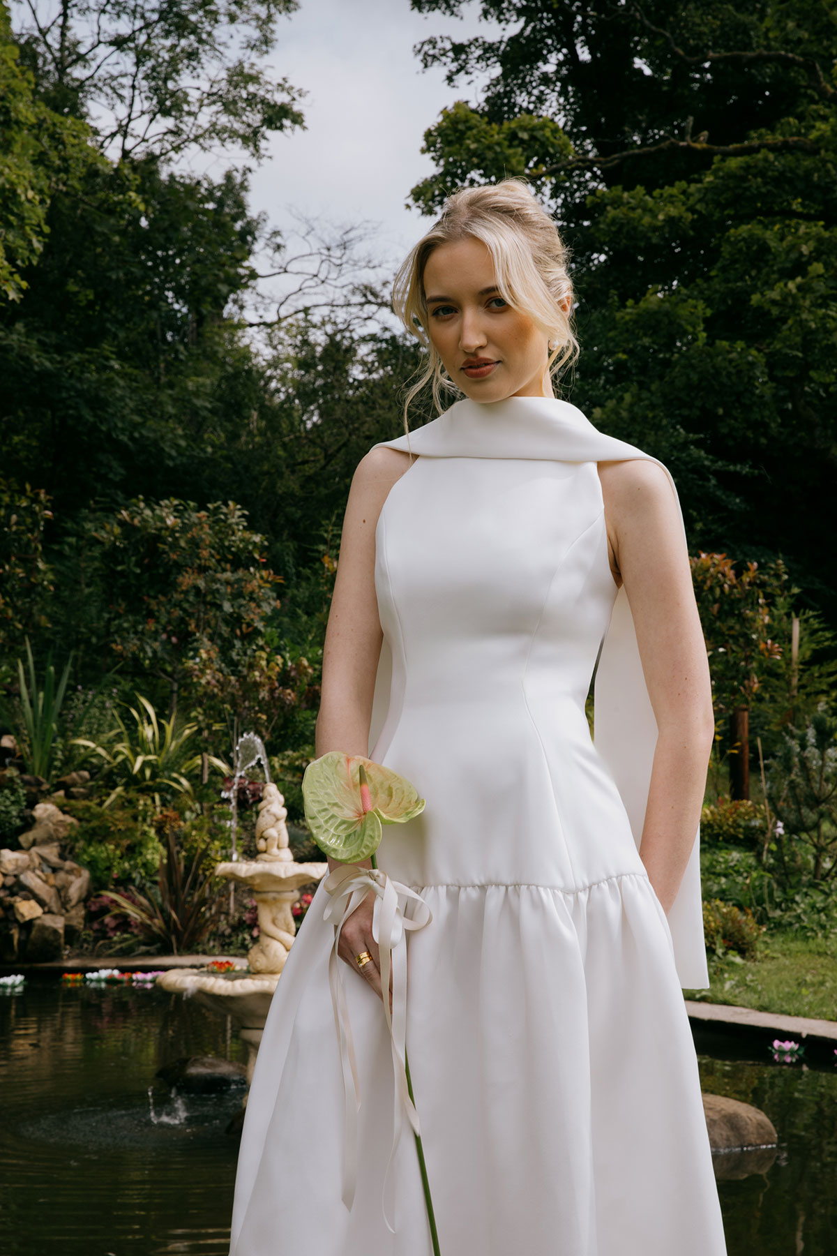 A bride in a sleeveless white dress stands beside a pond and fountain, holding a single anthurium stem, surrounded by garden plants and trees.