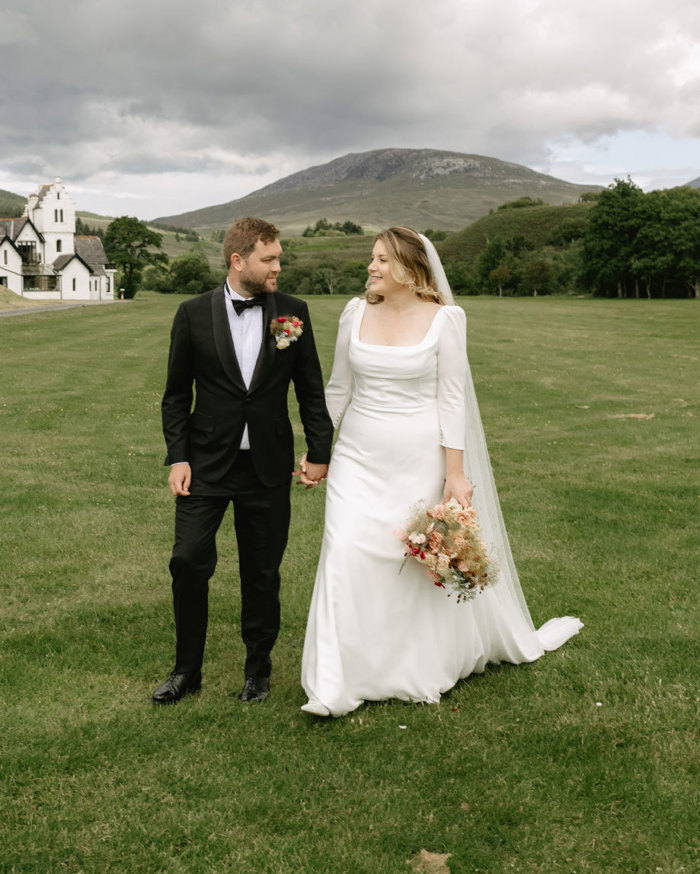 a bride and groom walking hand in hand across a lawn with white building and hills behind them