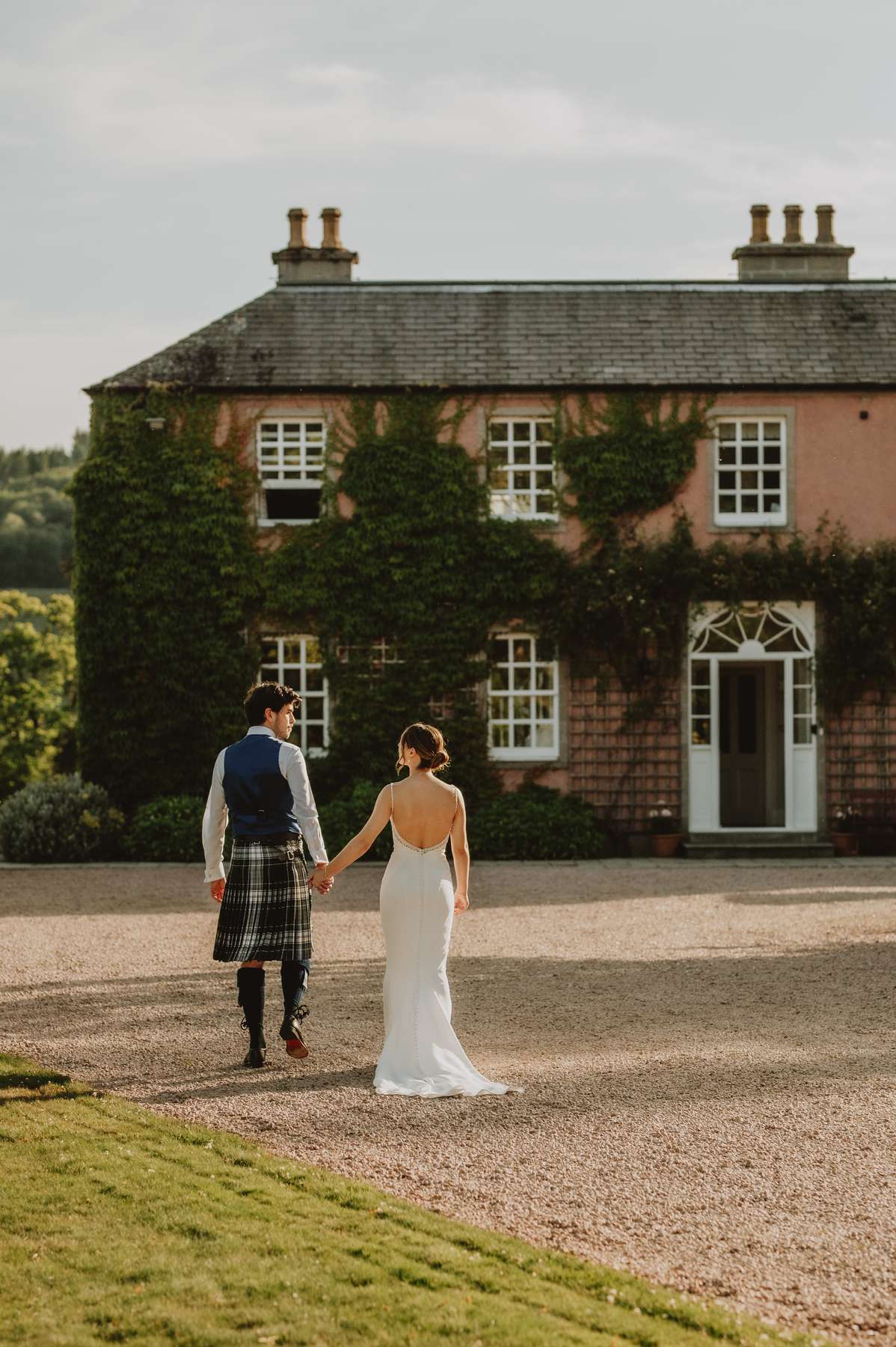 Newlyweds walk towards Ballogie House with its pink façade and ivy-covered walls behind them