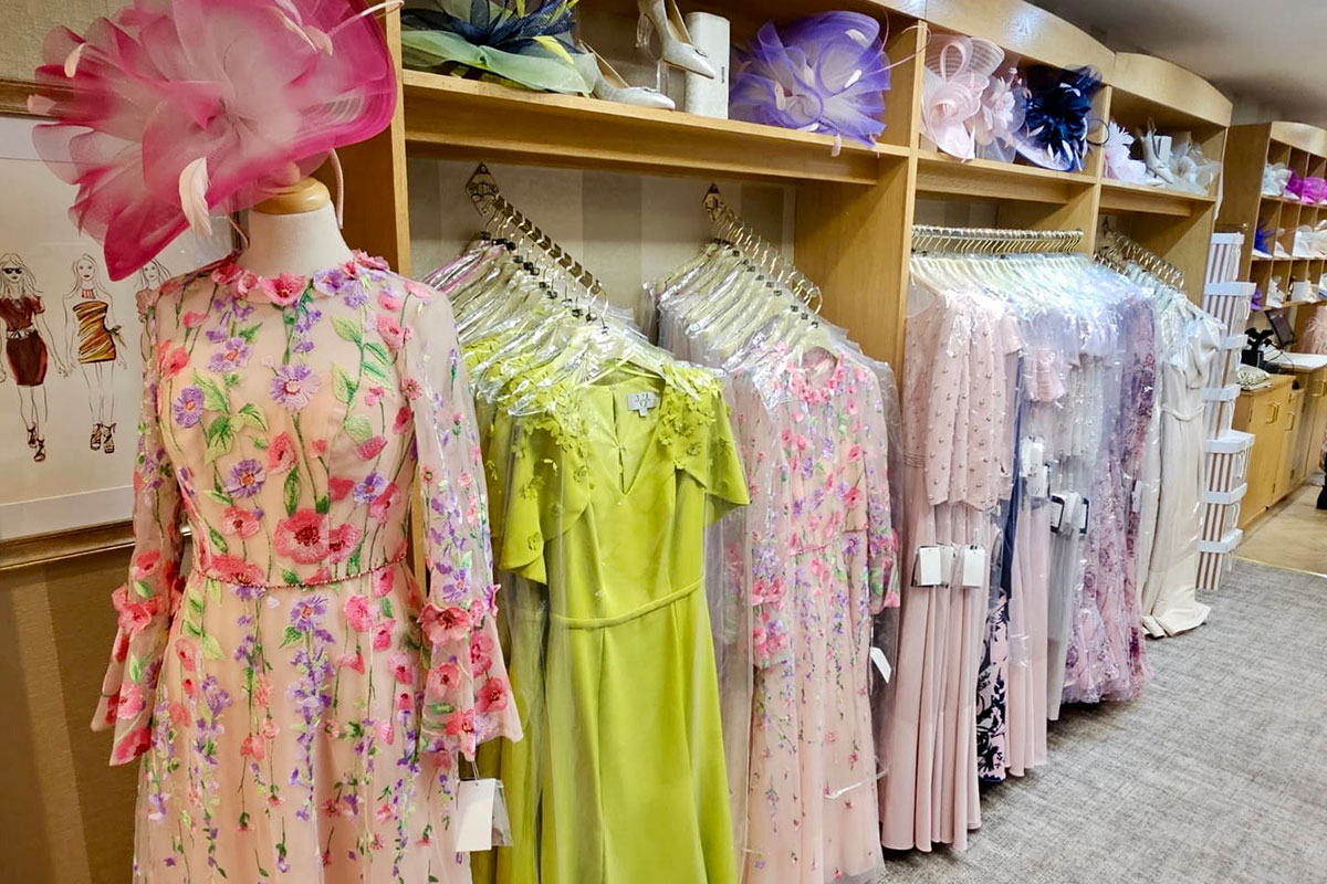 Interior of occasionwear boutique showing rails of pastel dresses and coordinating fascinators on shelves, with a mannequin in a floral embroidered dress and pink hat in the foreground