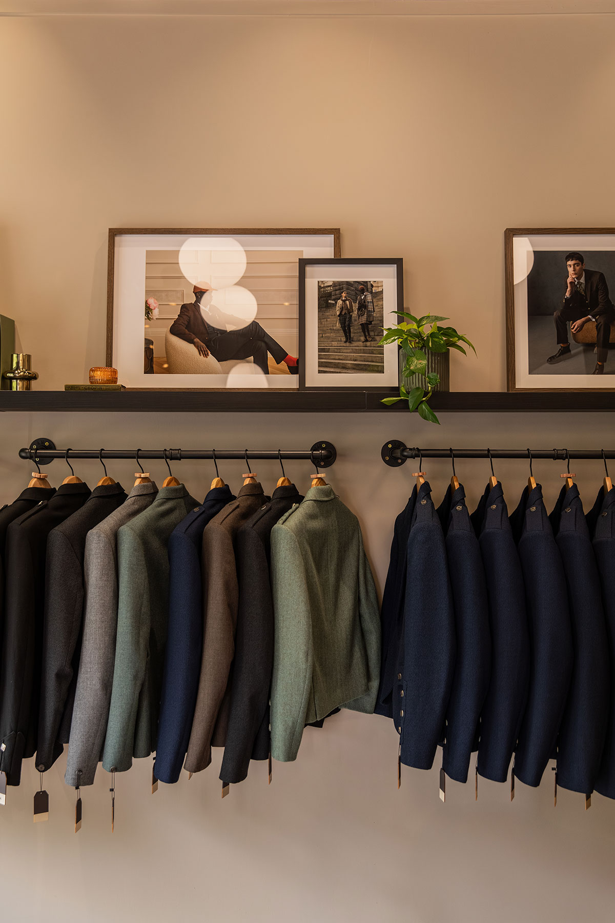 a wooden shelf with various sizes of photo frames and a plant resting on top above two hang rails of kilt outfit jackets hanging neatly with price tags hanging from the sleeves