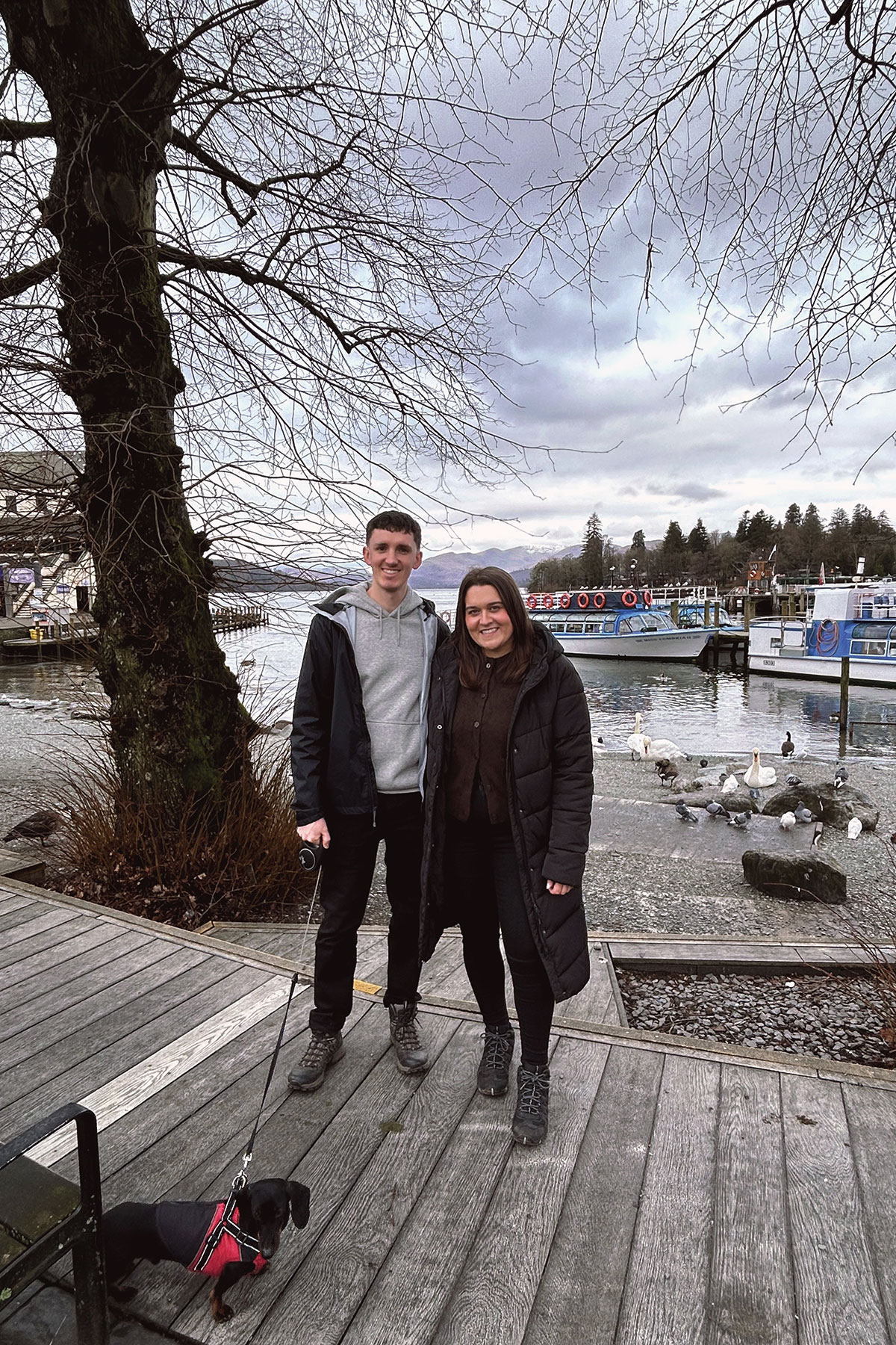 Couple standing by lakeside with boats and mountain views during engagement outing in Scotland