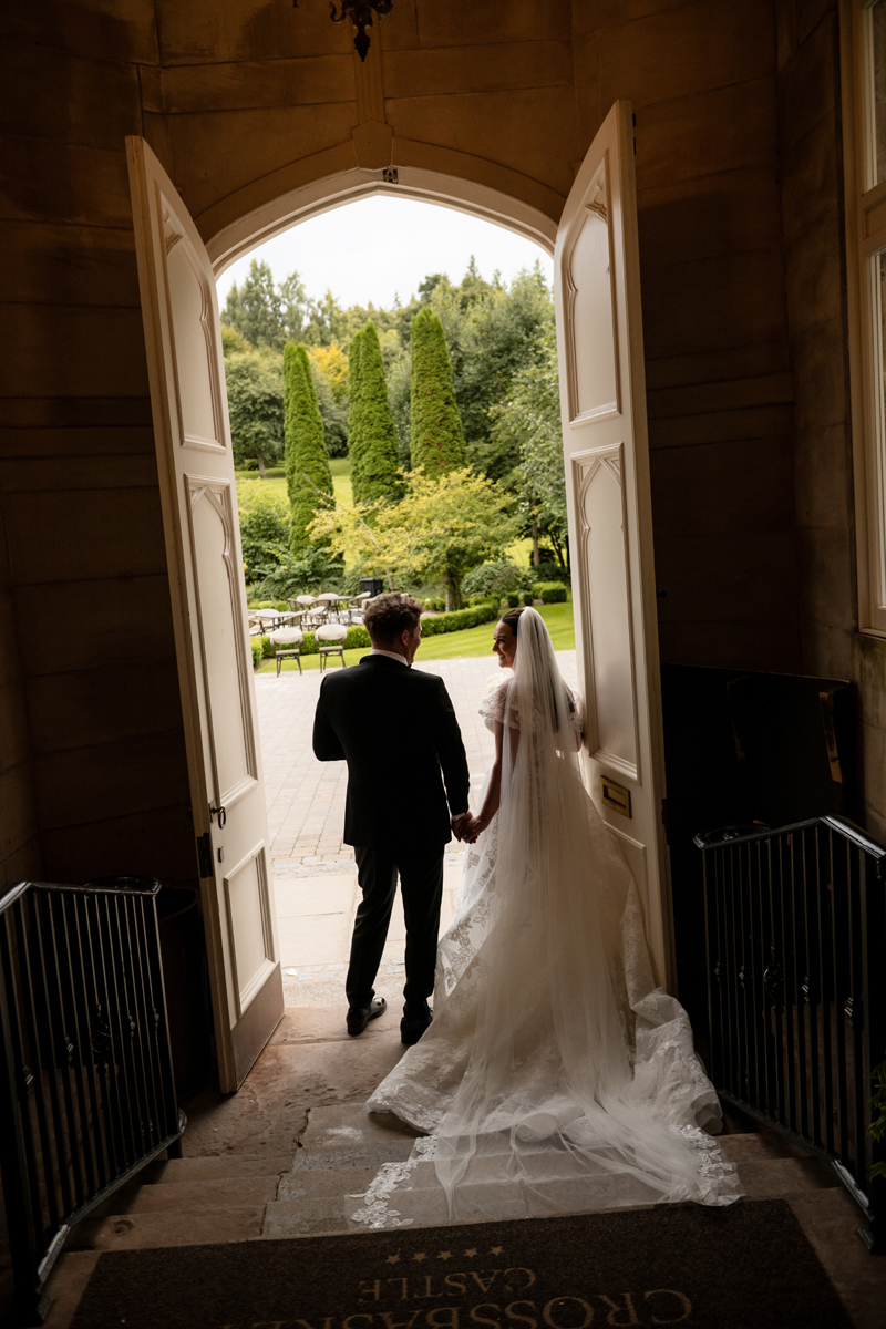 Bride and groom walking through the open doors of Crossbasket Castle towards the gardens on their wedding day.