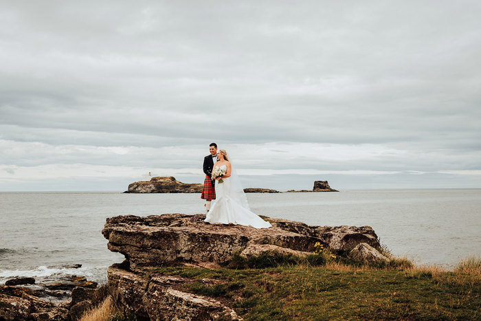 A groom in a kilt and a bride in wedding attire standing on a rock by water.