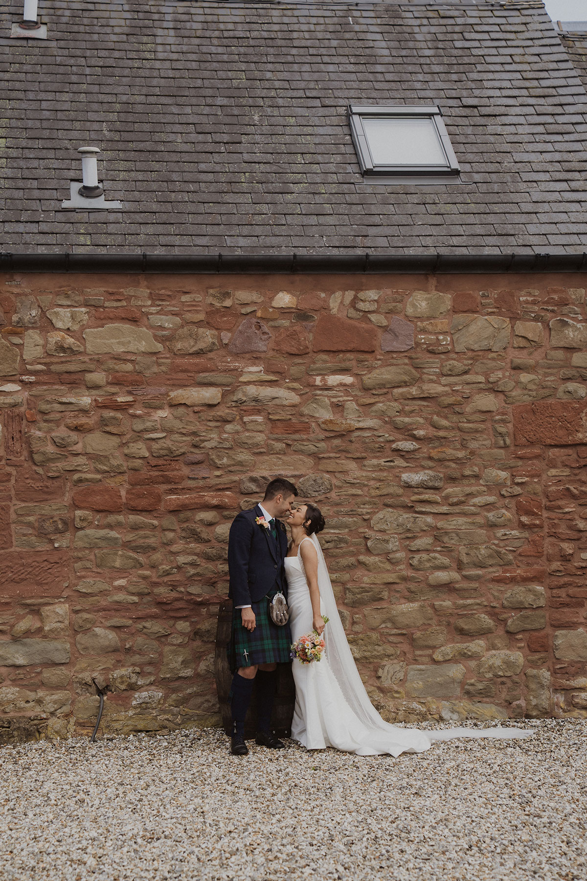 Bride and groom standing against rustic stone barn wall after countryside Scottish wedding ceremony
