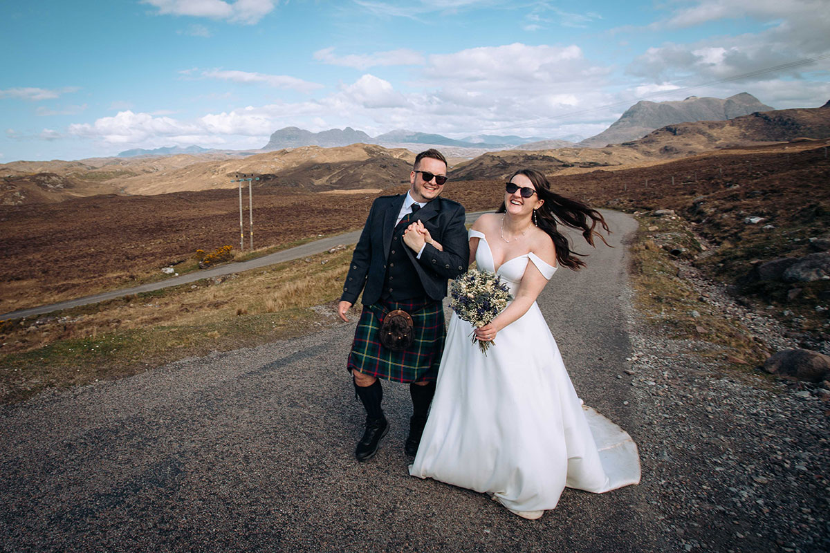Bride and groom walk hand in hand on a quiet Highland road surrounded by rugged mountain views near Achnahaird Bay. 