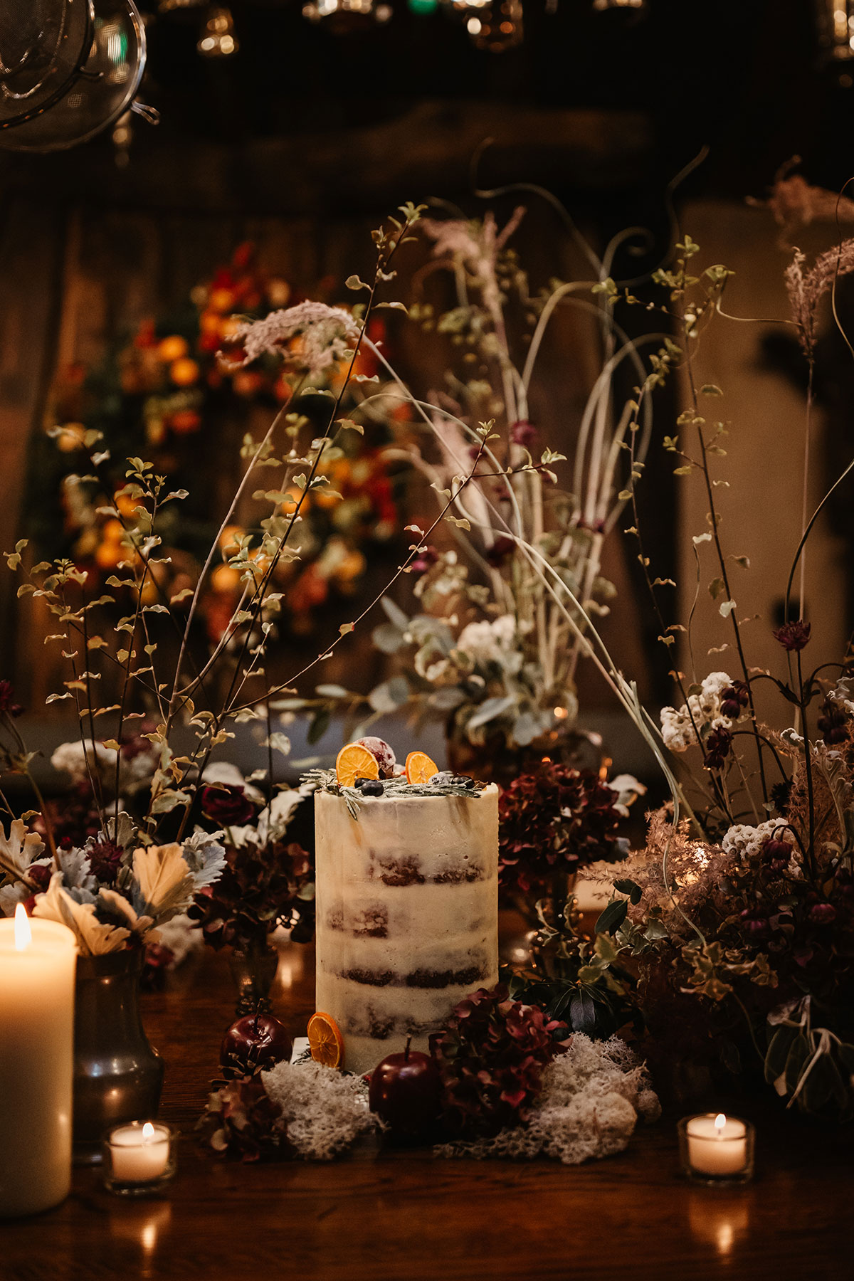 Tall semi-naked wedding cake decorated with citrus slices, surrounded by candlelight and winter florals on a rustic table