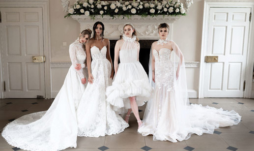 four women in bridalwear stand alongside each other in a horizontal line striking various poses to show off their dresses in front of an unused white marble fireplace decorated with white roses