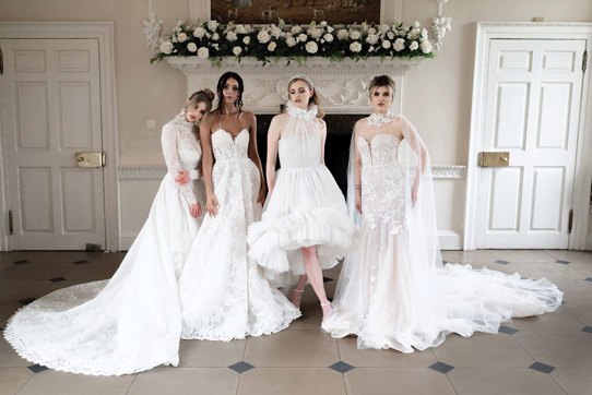 four women in bridalwear stand alongside each other in a horizontal line striking various poses to show off their dresses in front of an unused white marble fireplace decorated with white roses