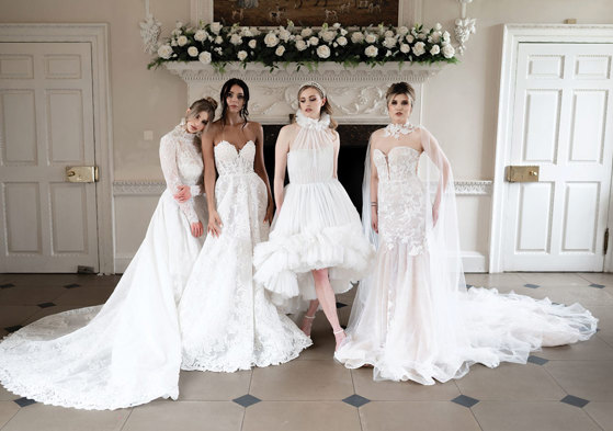 four women in bridalwear stand alongside each other in a horizontal line striking various poses to show off their dresses in front of an unused white marble fireplace decorated with white roses