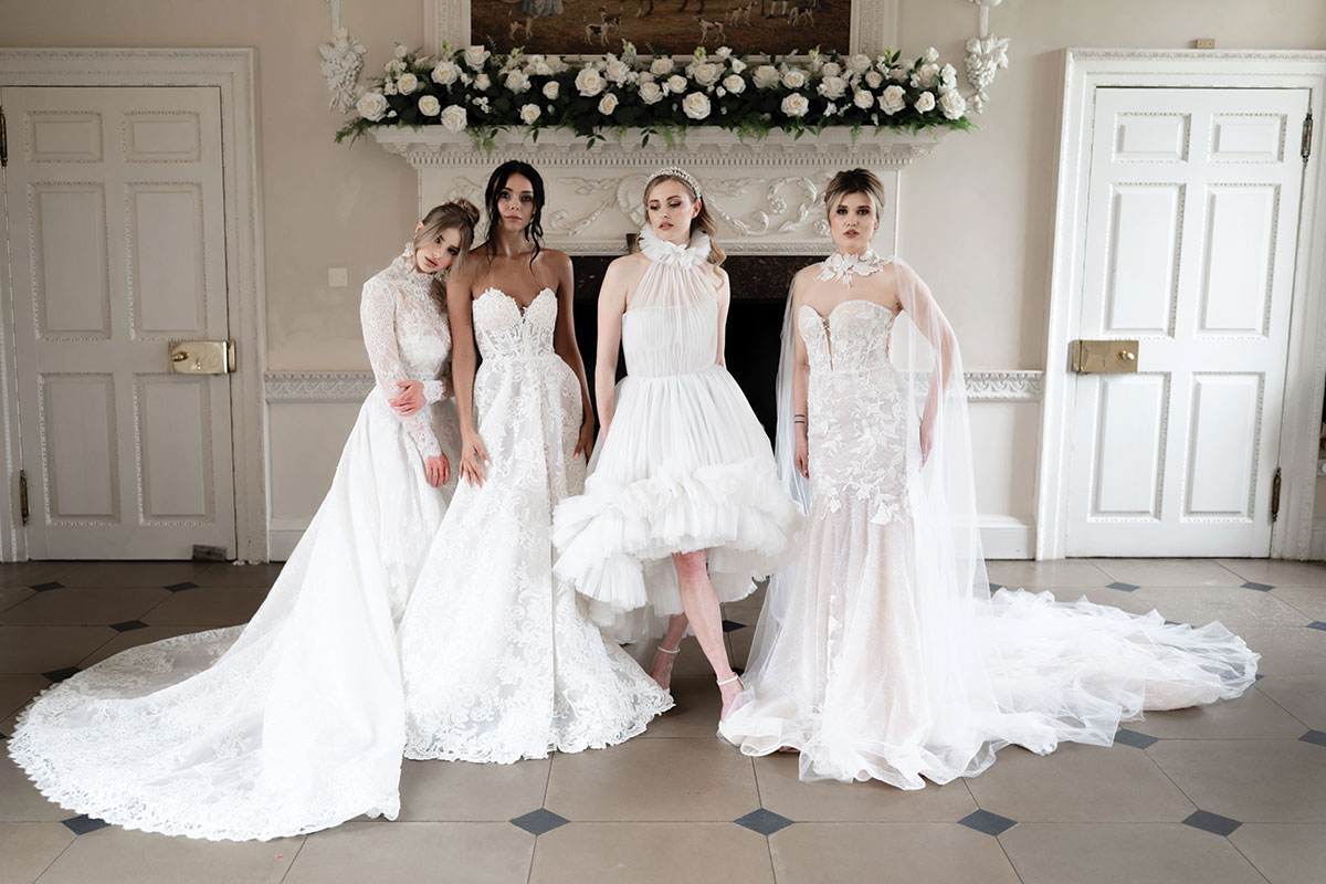 four women in bridalwear stand alongside each other in a horizontal line striking various poses to show off their dresses in front of an unused white marble fireplace decorated with white roses