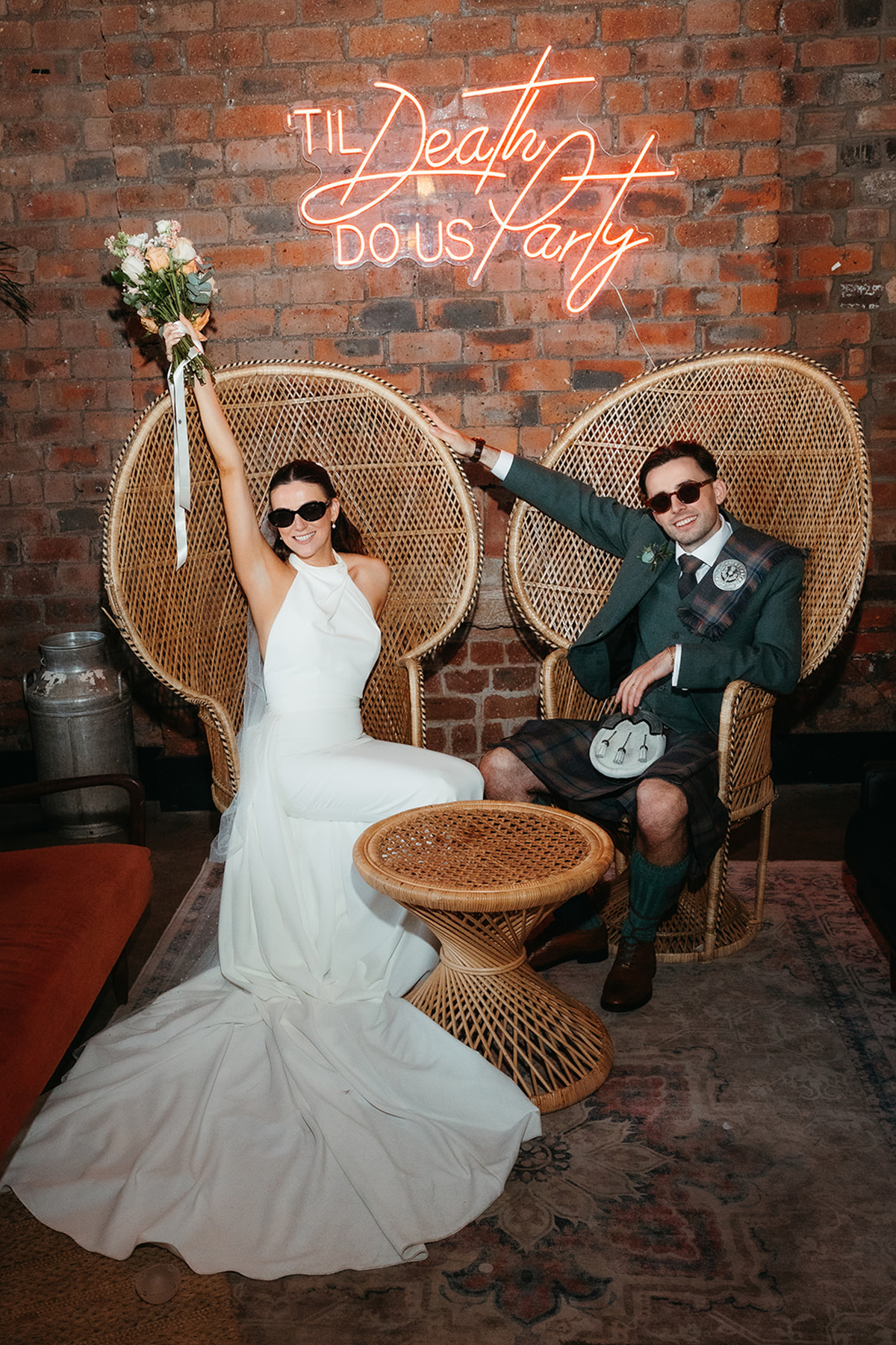 Bride and groom pose in wicker chairs beneath a neon sign at The Engine Works wedding venue