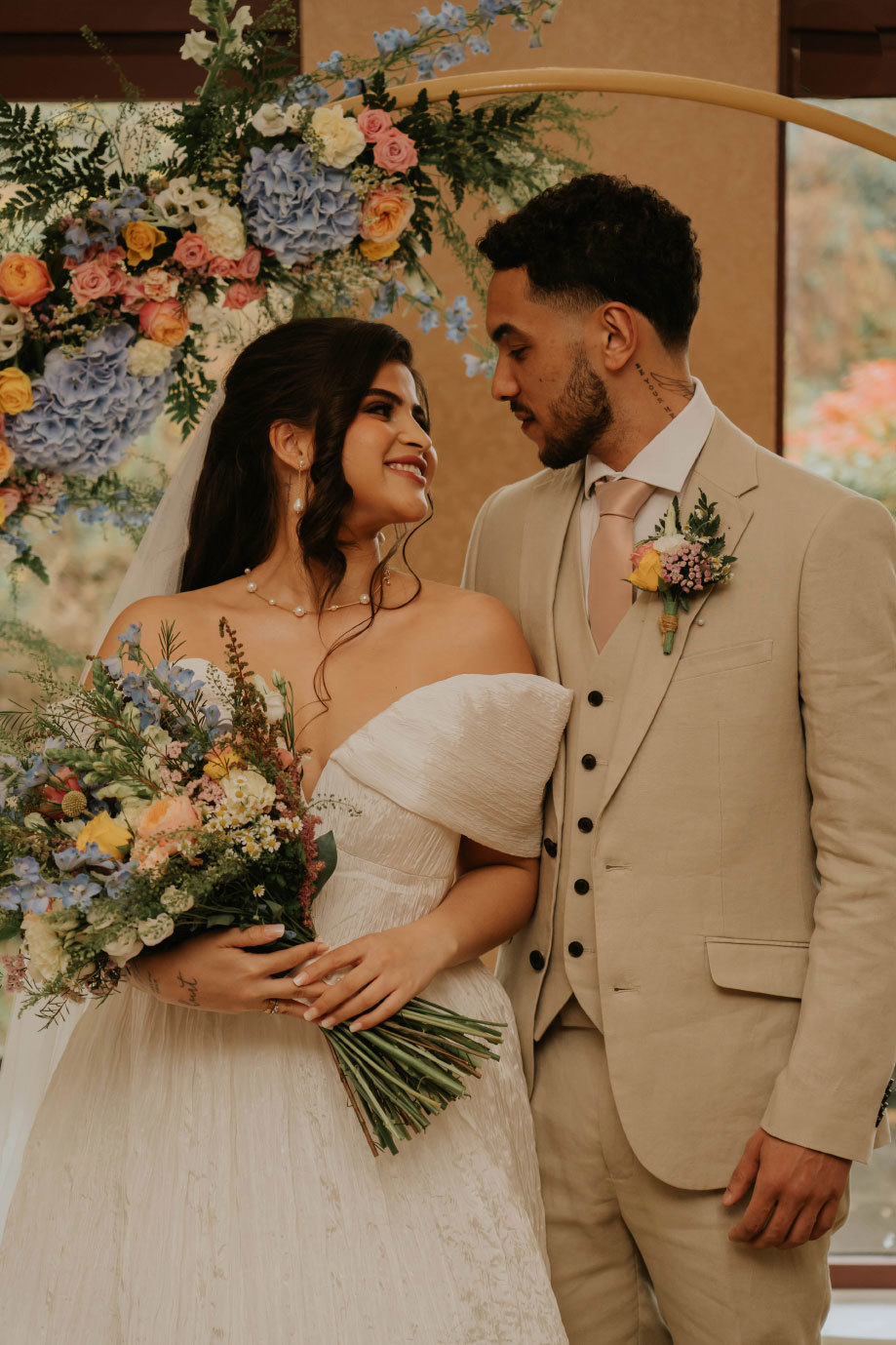 A bride and groom look lovingly at each other standing in front of a colourful flower arch