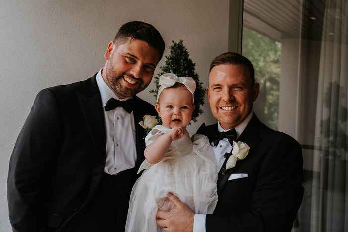 Two men wearing black tie suits posing with a baby.