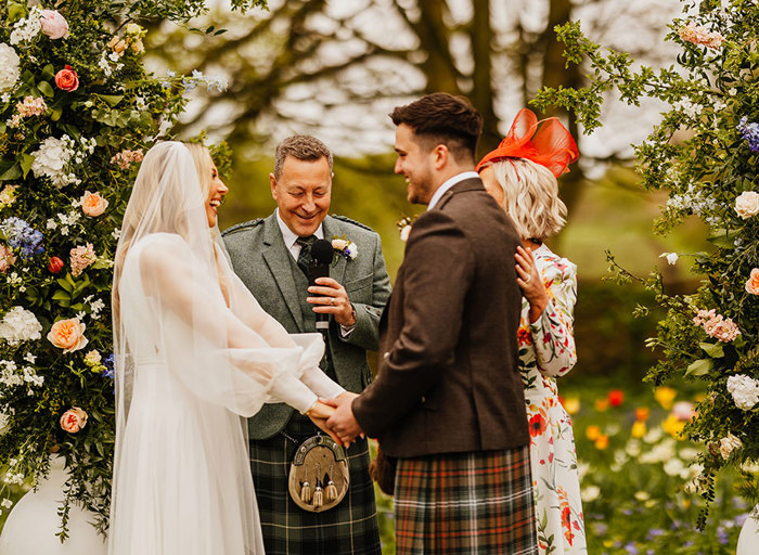 a bride and groom laughing during a wedding ceremony. They stand in front of a person wearing a kilt and a person wearing a floral patterned dress. They are in a flower filled garden setting with tall pastel floral arrangements framing either side