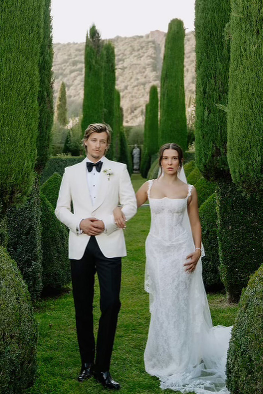 bride in lace corseted wedding dress and groom in white jacket tux stand arm in arm within green hedged garden