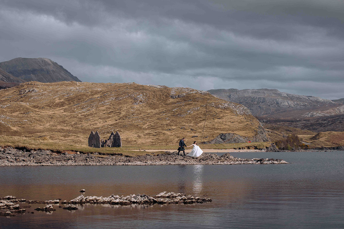 Bride and groom pose together by the water at Achnahaird Bay, with dramatic hills and coastal scenery behind them.