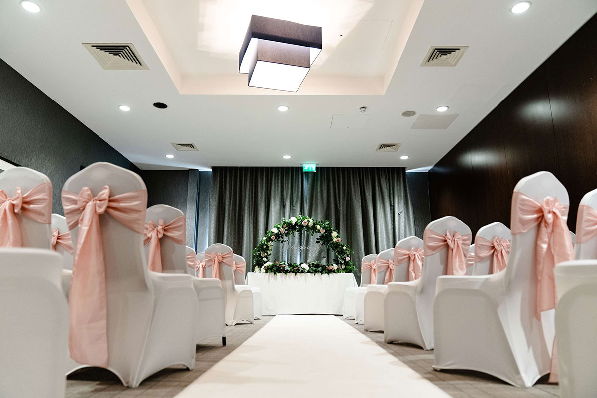 Wedding ceremony room dressed with blush chair sashes, floral ceremony arch and white aisle at a modern Scottish wedding venue