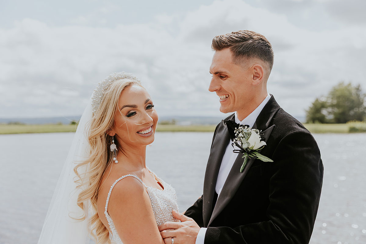 blonde bride and groom in tux stand across from each other smiling with body of water behind them