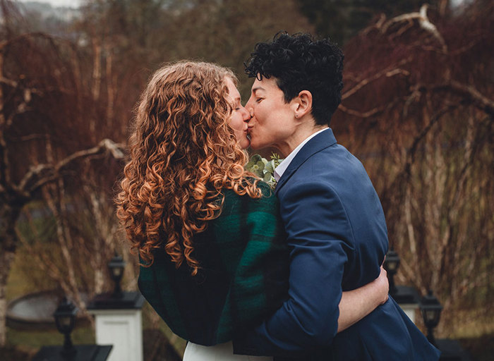 Two women kissing wearing a blue suit and a green tartan shawl