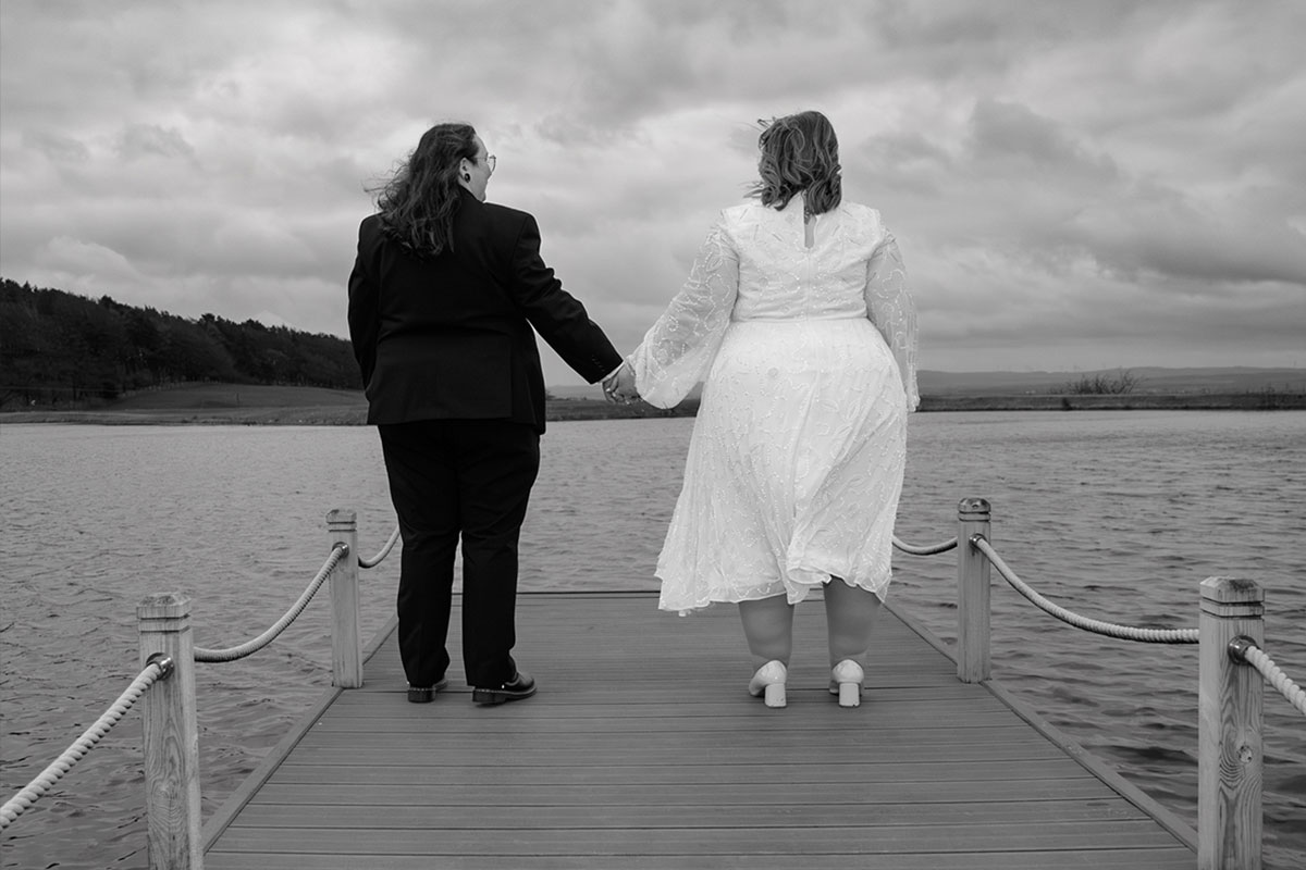 A couple stand holding hands at the end of a pier with water and hills in the distance