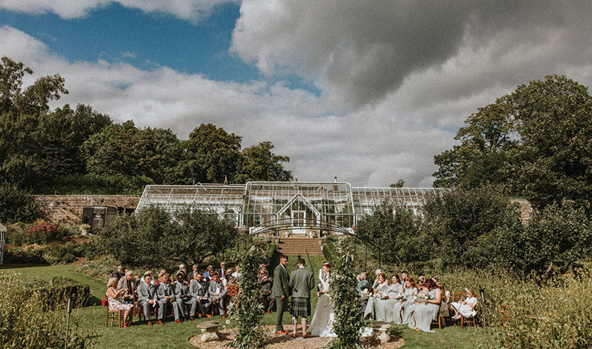 a group of people sitting on chairs watching a wedding ceremony in the garden of Teasses Estate with Victorian glasshouse in background