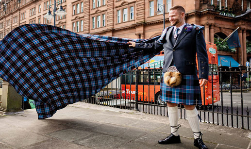 Groom in blue tartan kilt outfit holds his arm out to support matching tartan train 