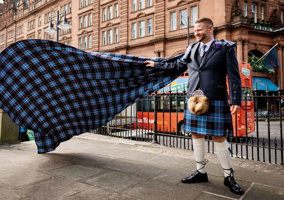 Groom in blue tartan kilt outfit holds his arm out to support matching tartan train 