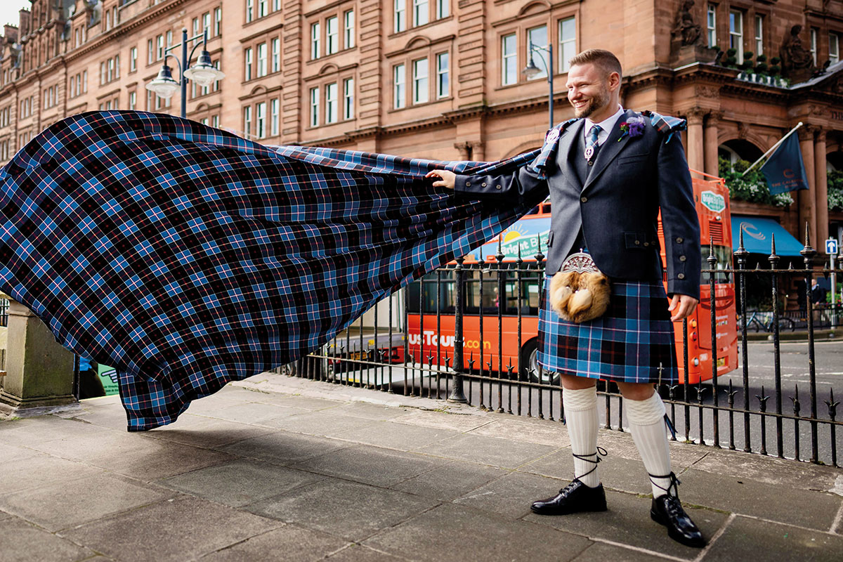Groom in blue tartan kilt outfit holds his arm out to support matching tartan train