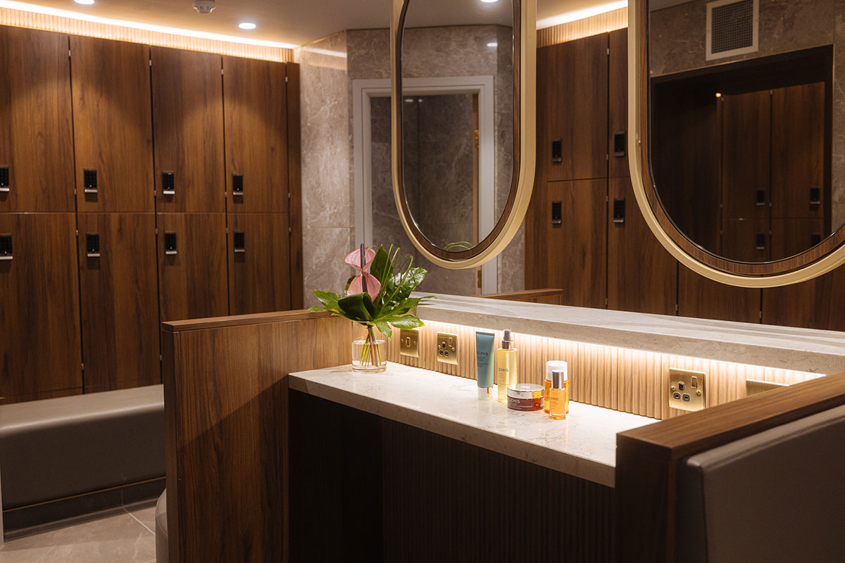 Well-lit changing room with wood-panelled lockers, marble walls, circular mirrors, and skincare products on a countertop