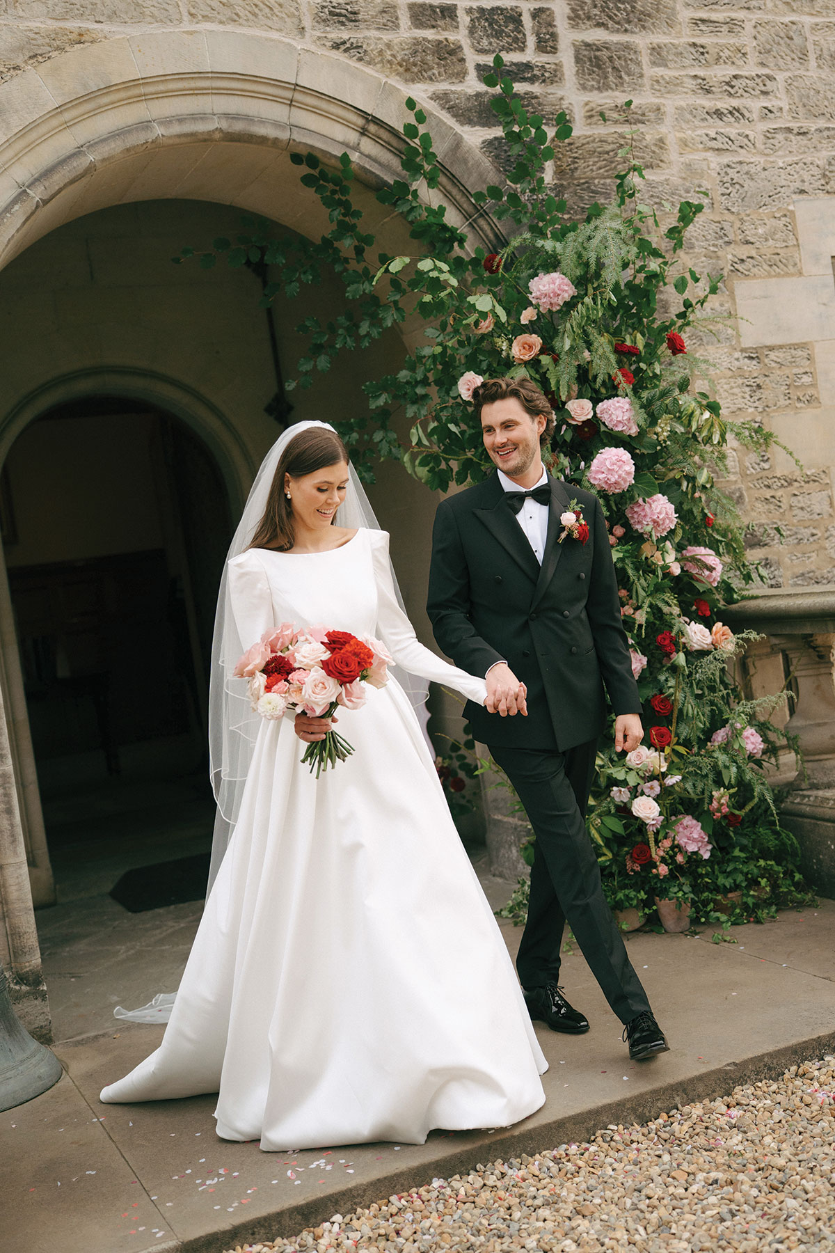 Bride Nicole and groom Ryan walk hand in hand outside Barnbougle Castle in Edinburgh, beside a floral display of pink and red roses.