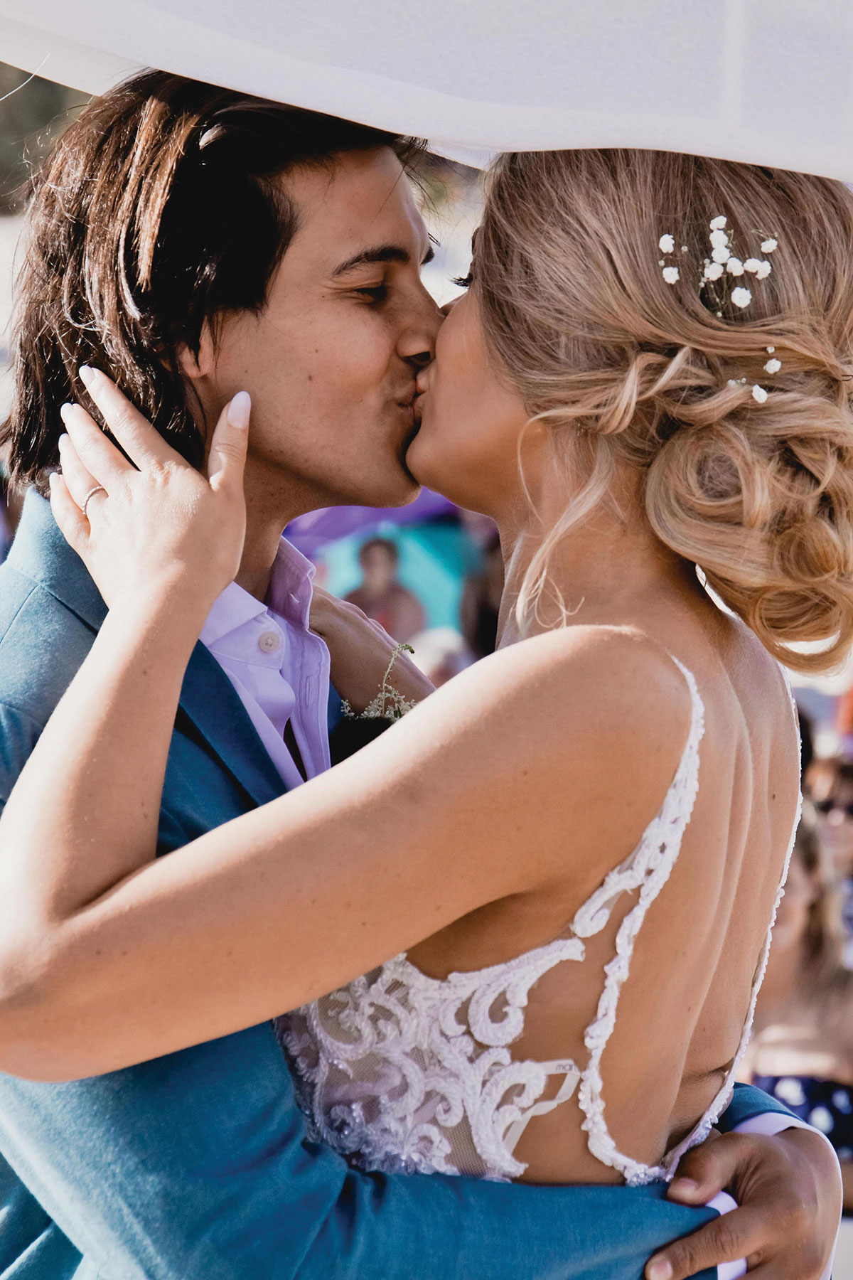 Bride and groom sharing a kiss outdoors; the bride wears a lace-back wedding dress with a soft updo decorated with small white flowers, and the groom wears a blue suit