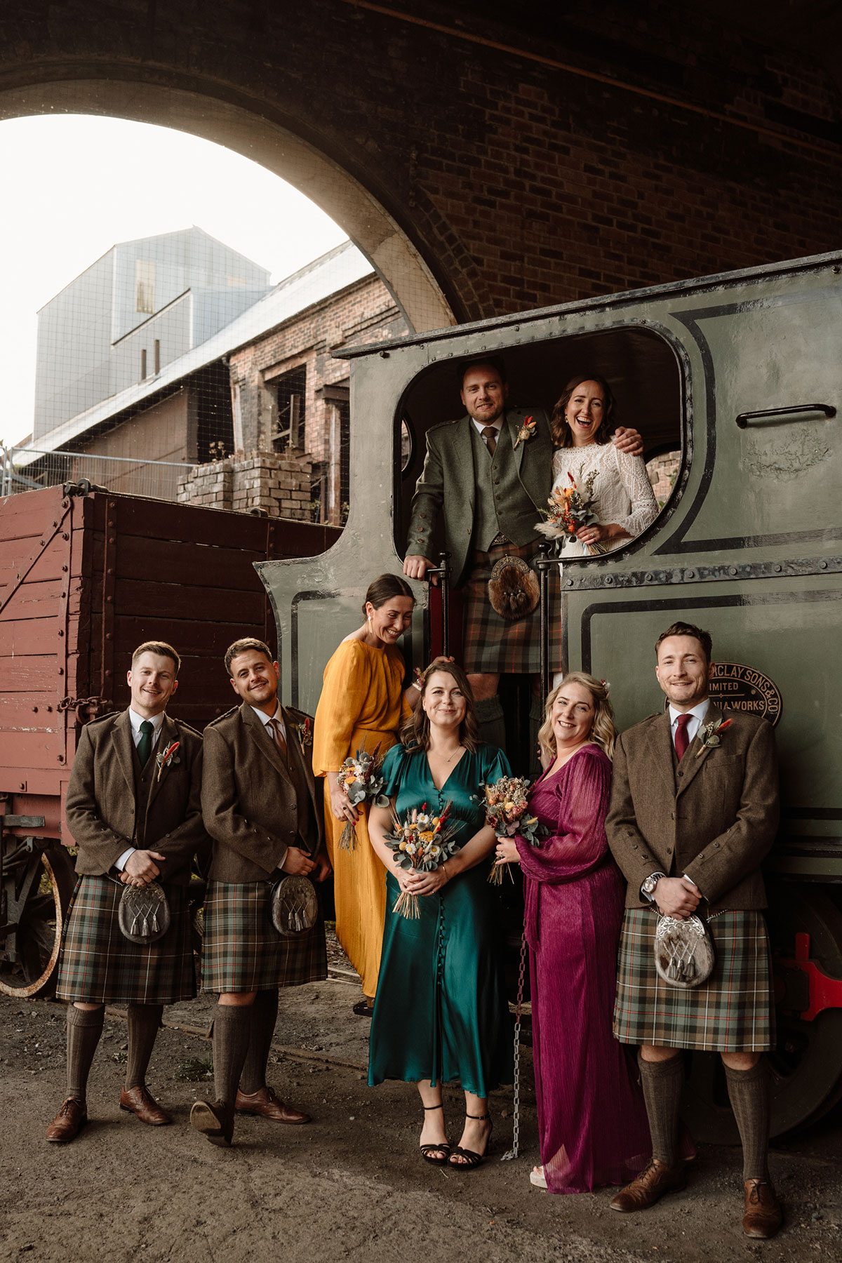 Bride, groom and wedding party gathered around a vintage steam train at an industrial heritage wedding venue