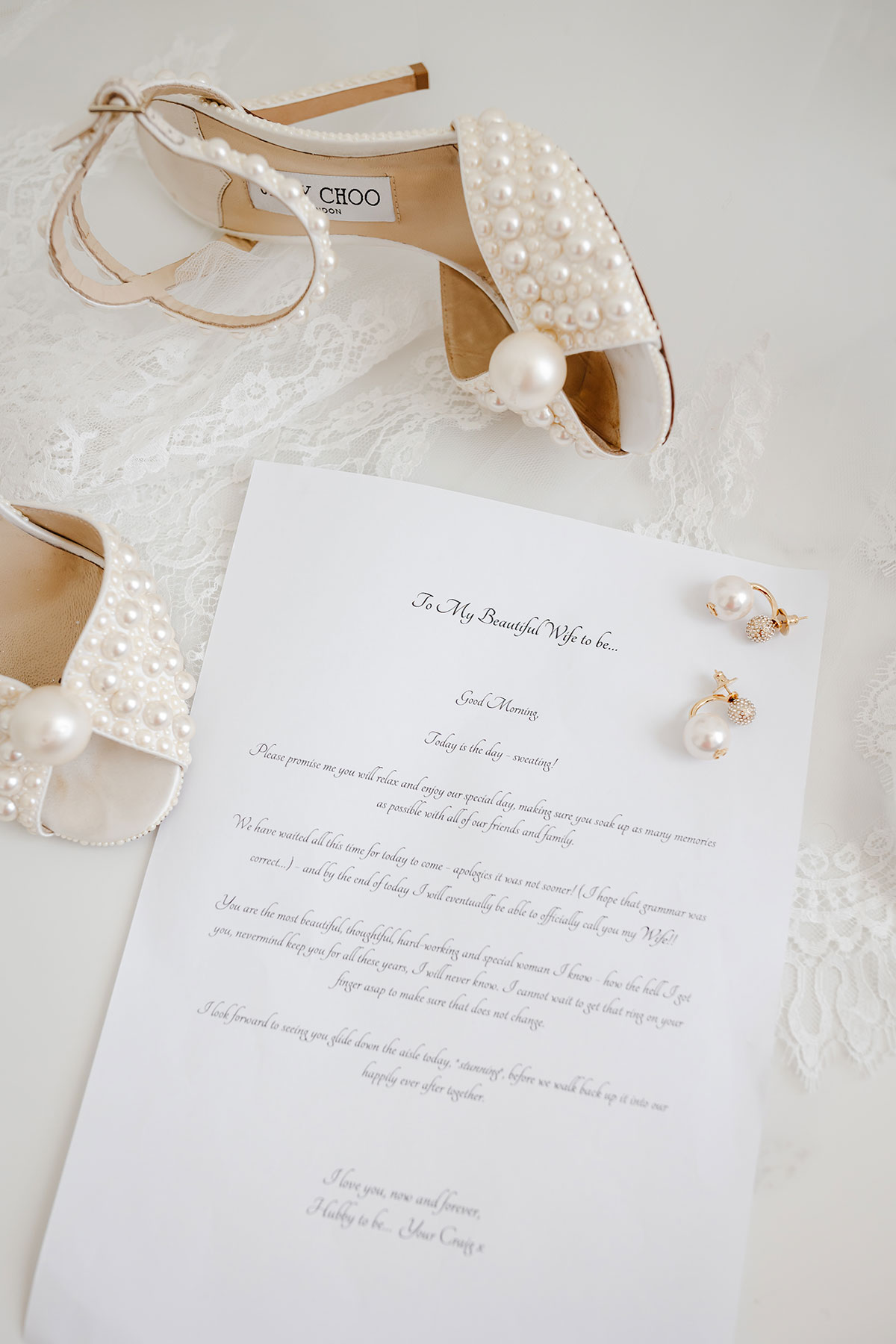 Pearl-embellished Jimmy Choo heels and pearl earrings beside a handwritten wedding letter on lace fabric