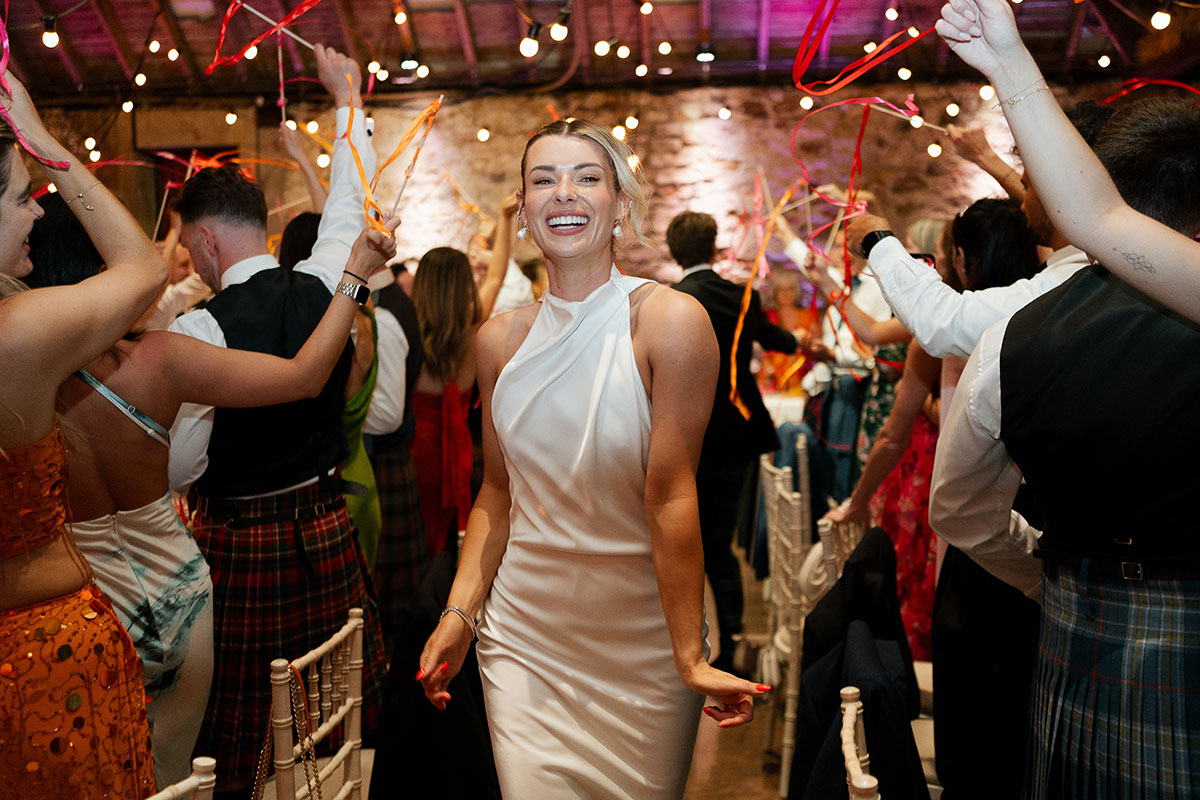 Bride dances into the wedding breakfast surrounded by guests waving colourful ribbon streamers inside Kinkell Byre's rustic barn reception space.