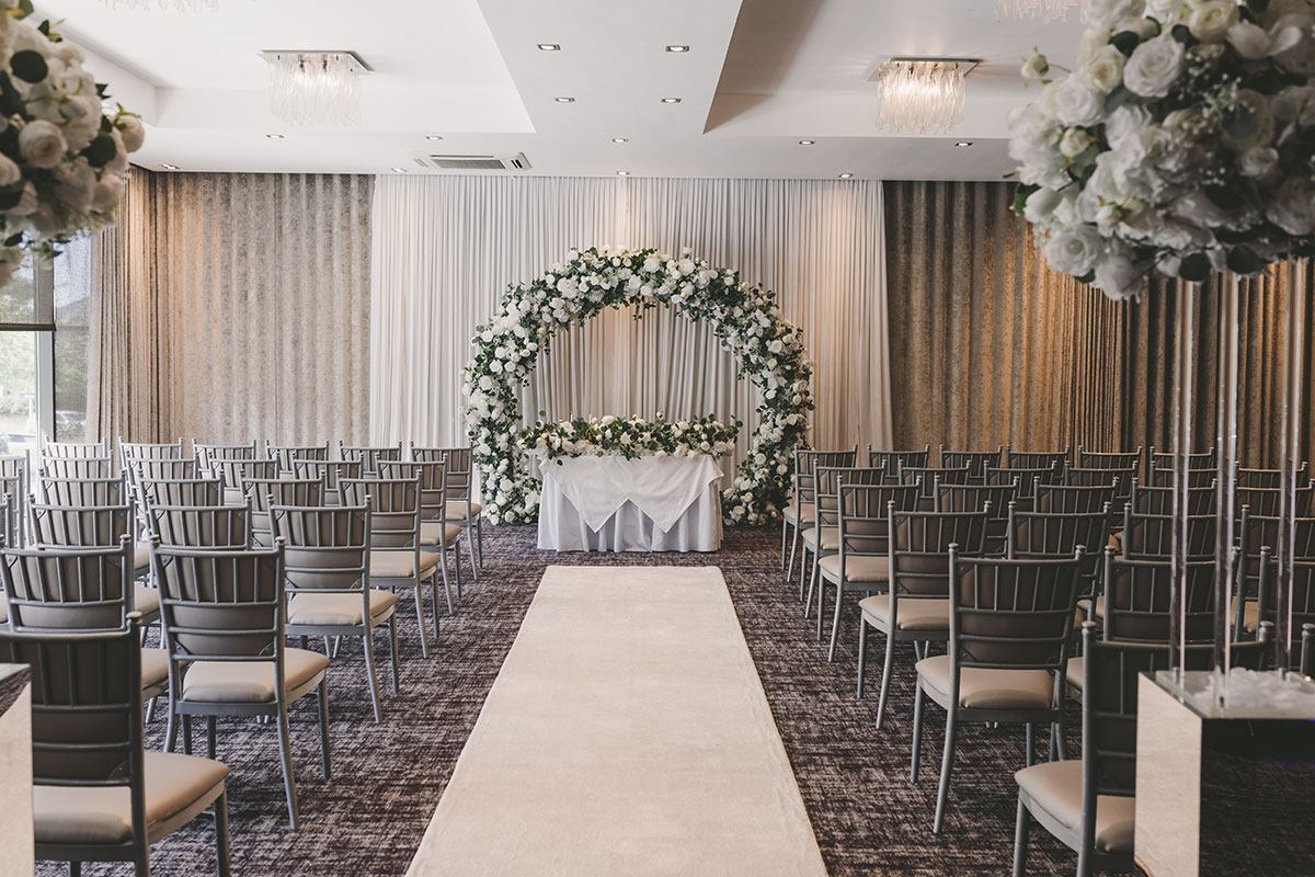Indoor wedding ceremony setup at The Carlton Hotel with Chiavari chairs, floral arch and draped backdrop.