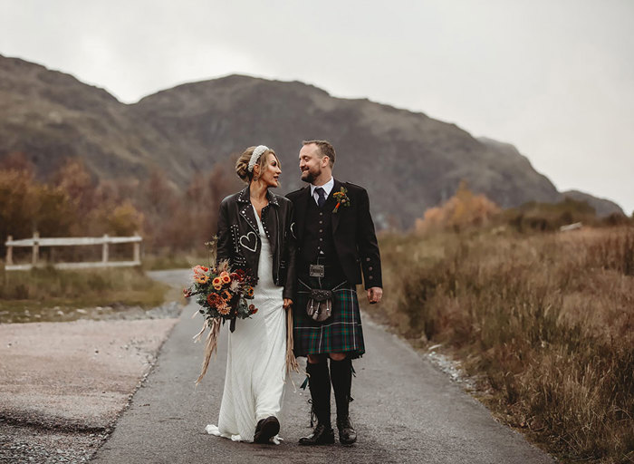 a bride wearing a black leather fringed jacket over her wedding dress walking with groom in a kilt in Perthshire countryside