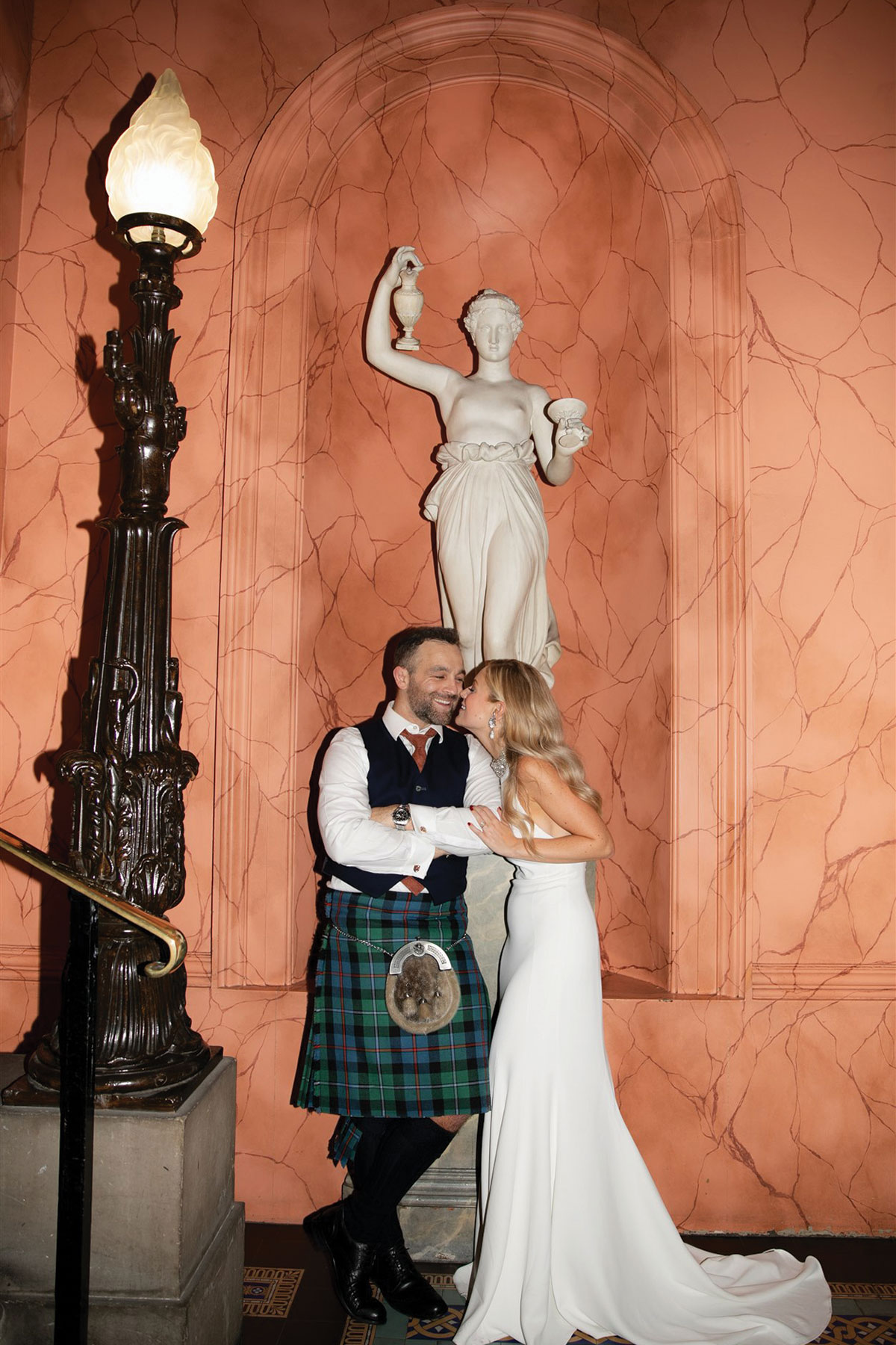 A bride and groom share a joyful moment beneath a marble statue and warm terracotta wall at their wedding venue