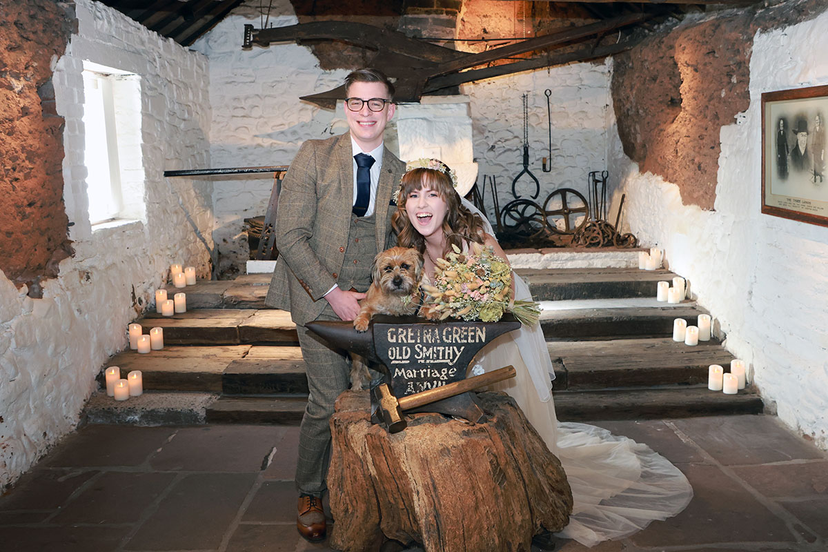 couple hold border terrier dog at gretna green ceremony room with wooden steps and candles in the background