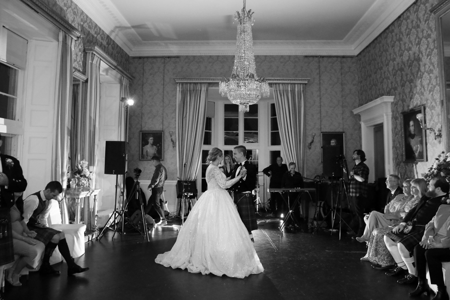A black and white photo of a bride and a groom dancing in a ballroom with their guests looking on 