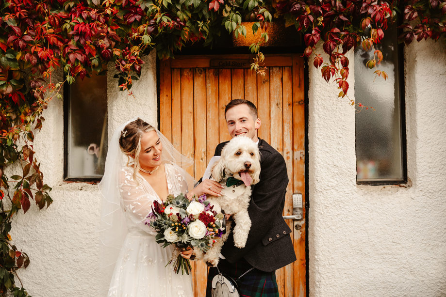 A bride and groom stand holding a small white dog and smiling 