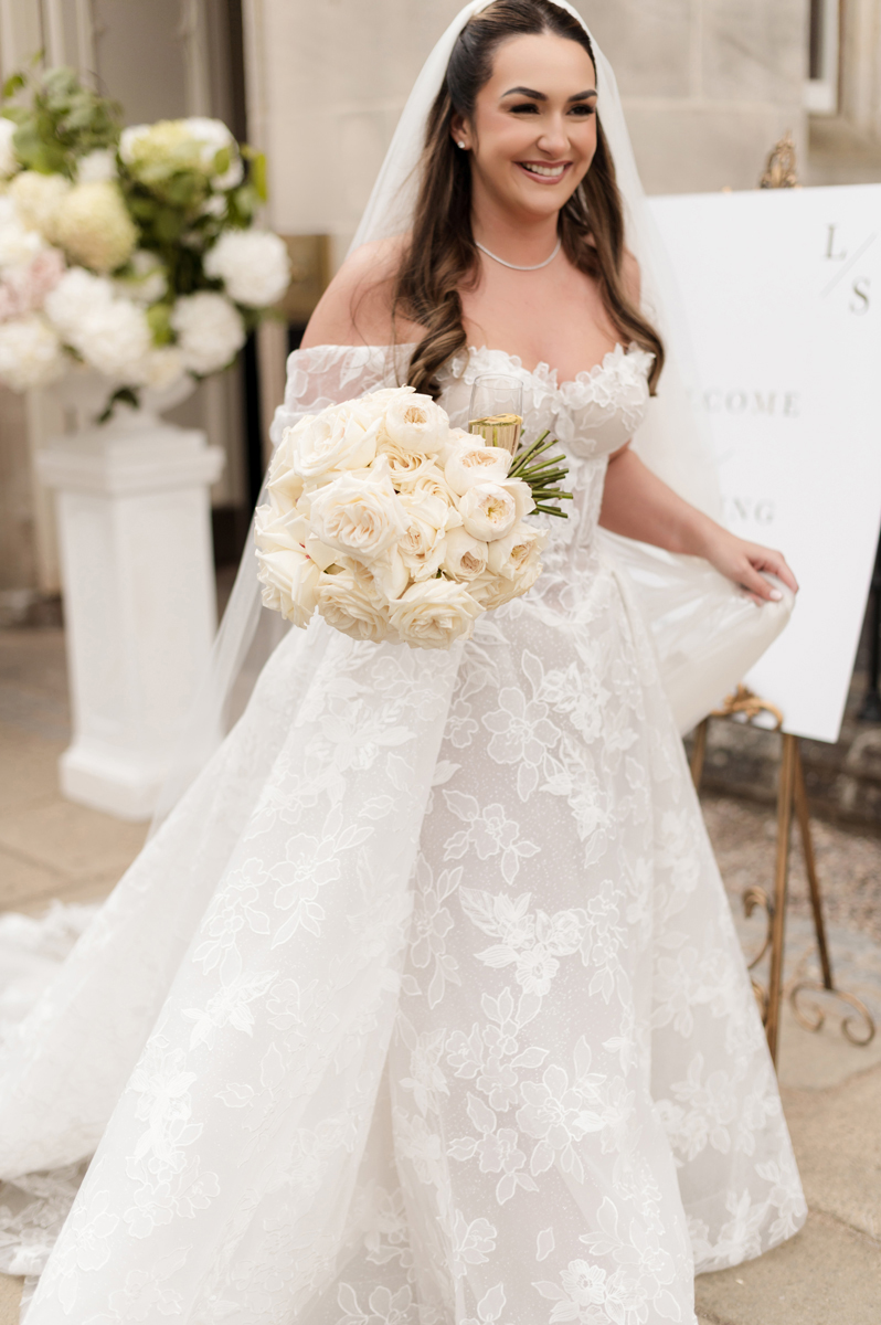 Smiling bride in an off-the-shoulder lace Galia Lahav wedding dress holding a bouquet of white roses at Crossbasket Castle.